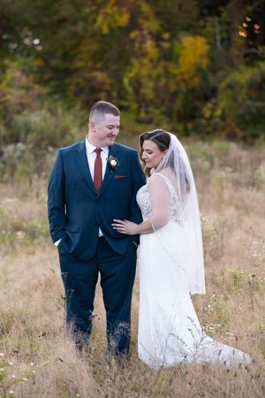 Bride and groom share tender moment in golden autumn field, groom in navy suit with burgundy tie.