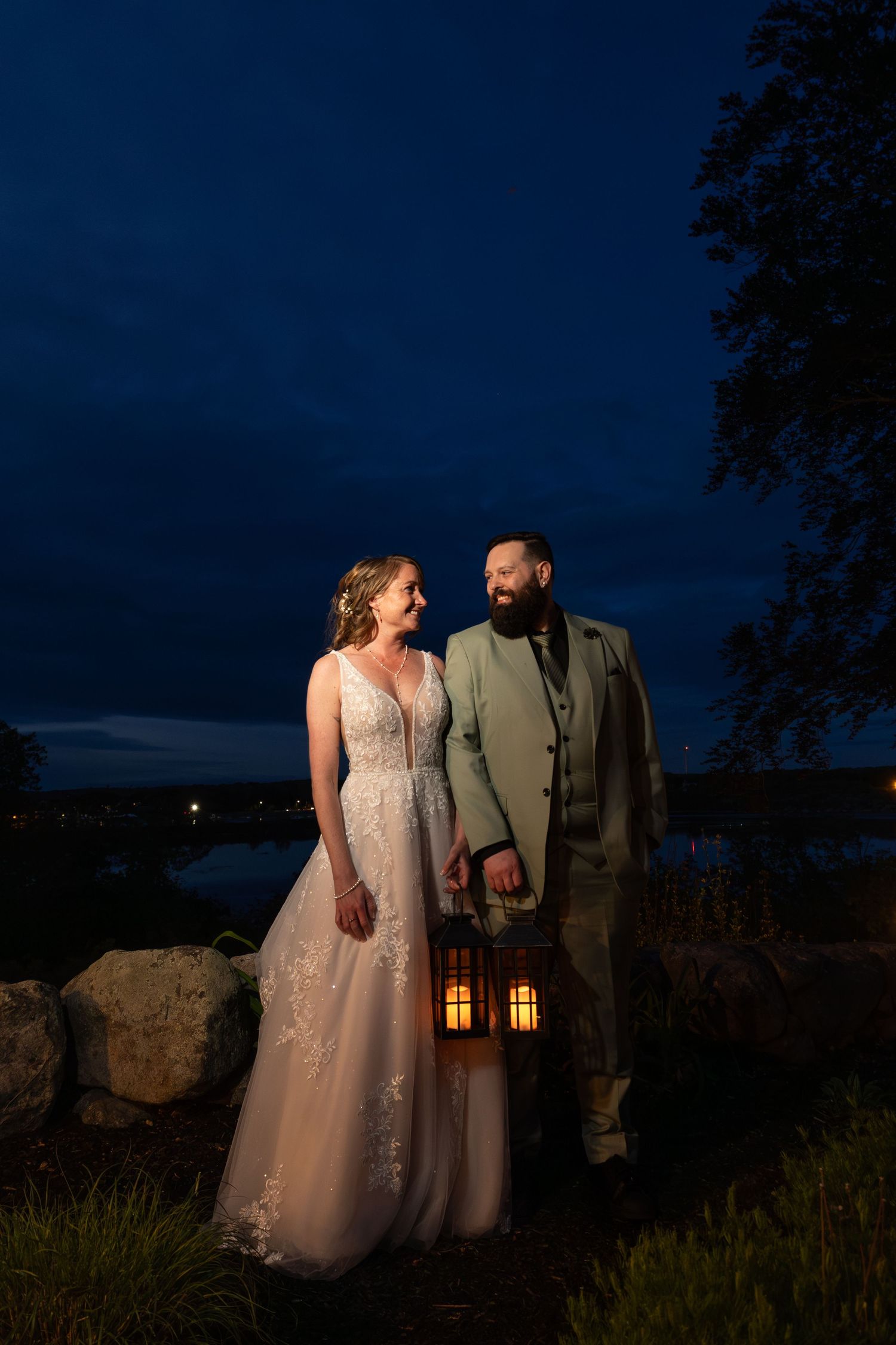 Bride and groom stand together at dusk holding a glowing lantern with a lake and city lights in the background.