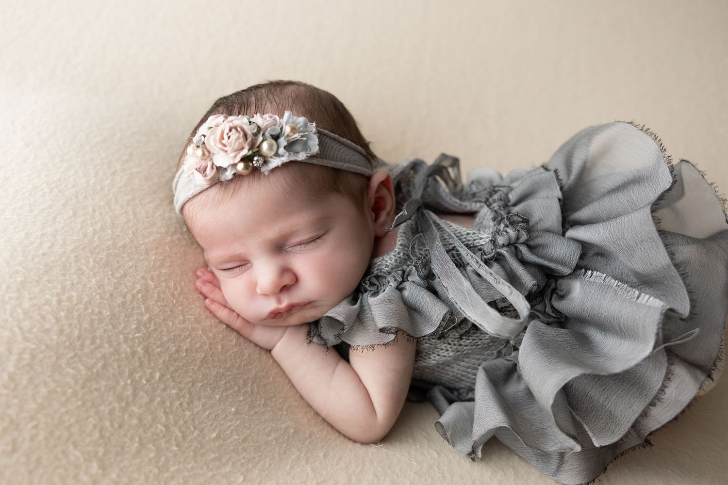 Sleeping newborn baby in gray ruffled outfit and floral headband on cream background.