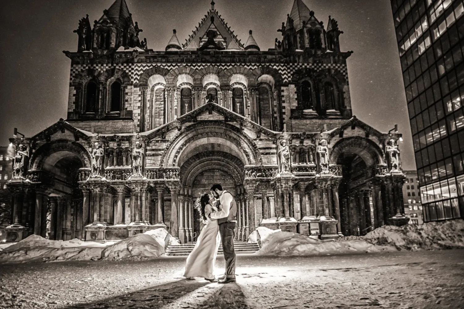 Black and white wedding photo of couple kissing in front of ornate Gothic cathedral in winter snow at night.