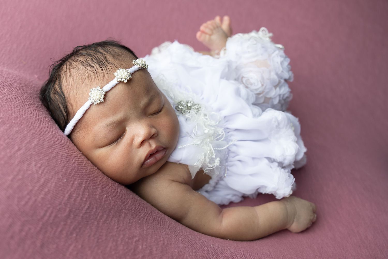 Sleeping newborn baby girl in white ruffled dress and pearl headband on pink background.