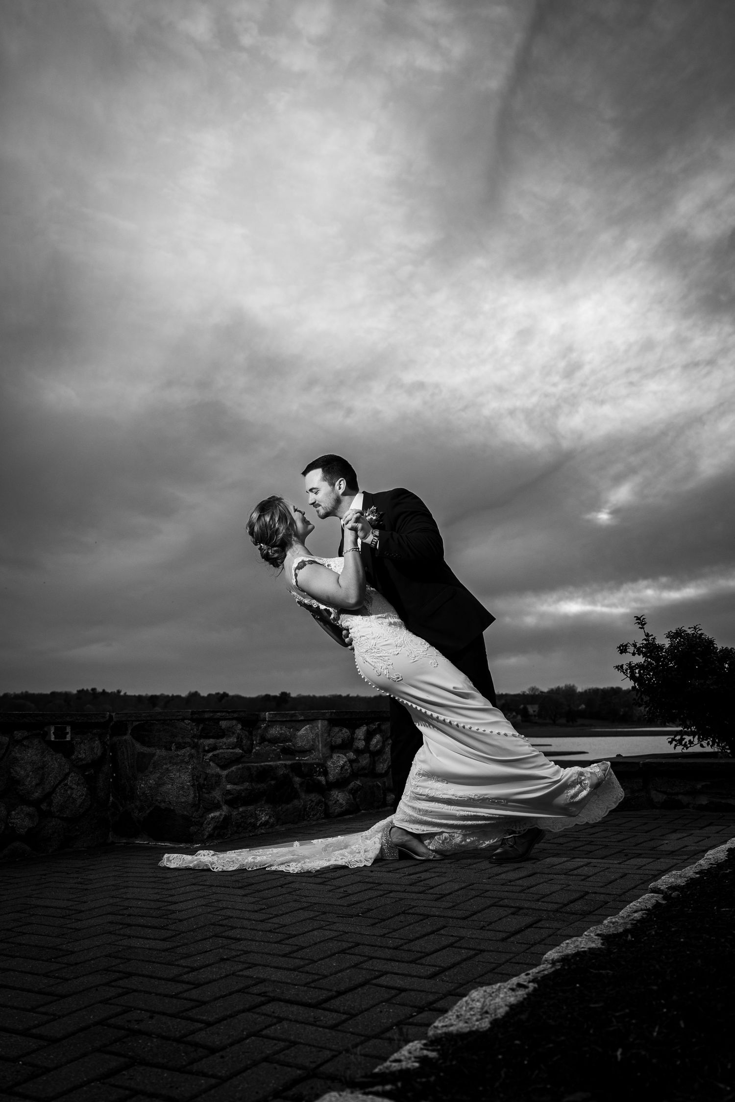 Black and white photo of a groom dipping his bride for a kiss outdoors under a dramatic cloudy sky.