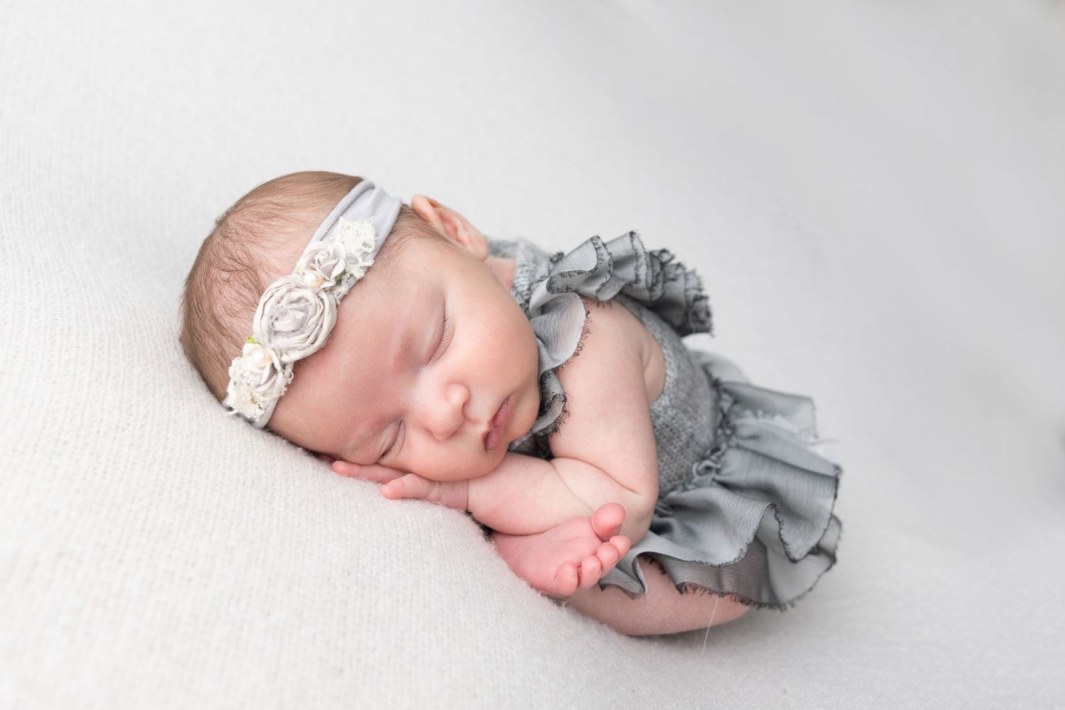 Peaceful newborn baby girl sleeping in gray ruffled romper and white floral headband on soft white blanket.