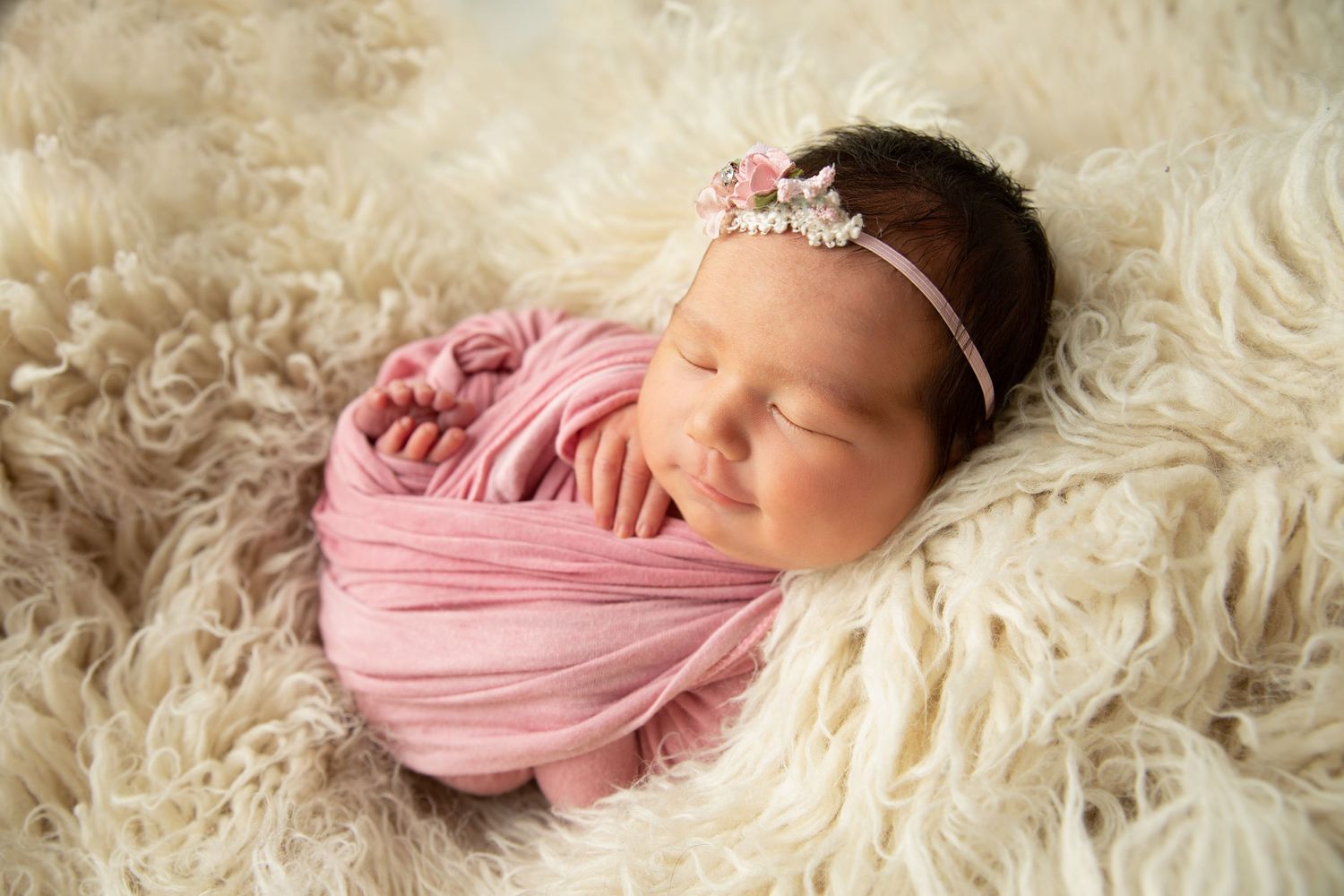 Newborn baby girl swaddled in pink wrap with floral headband sleeping on fluffy white fur blanket.
