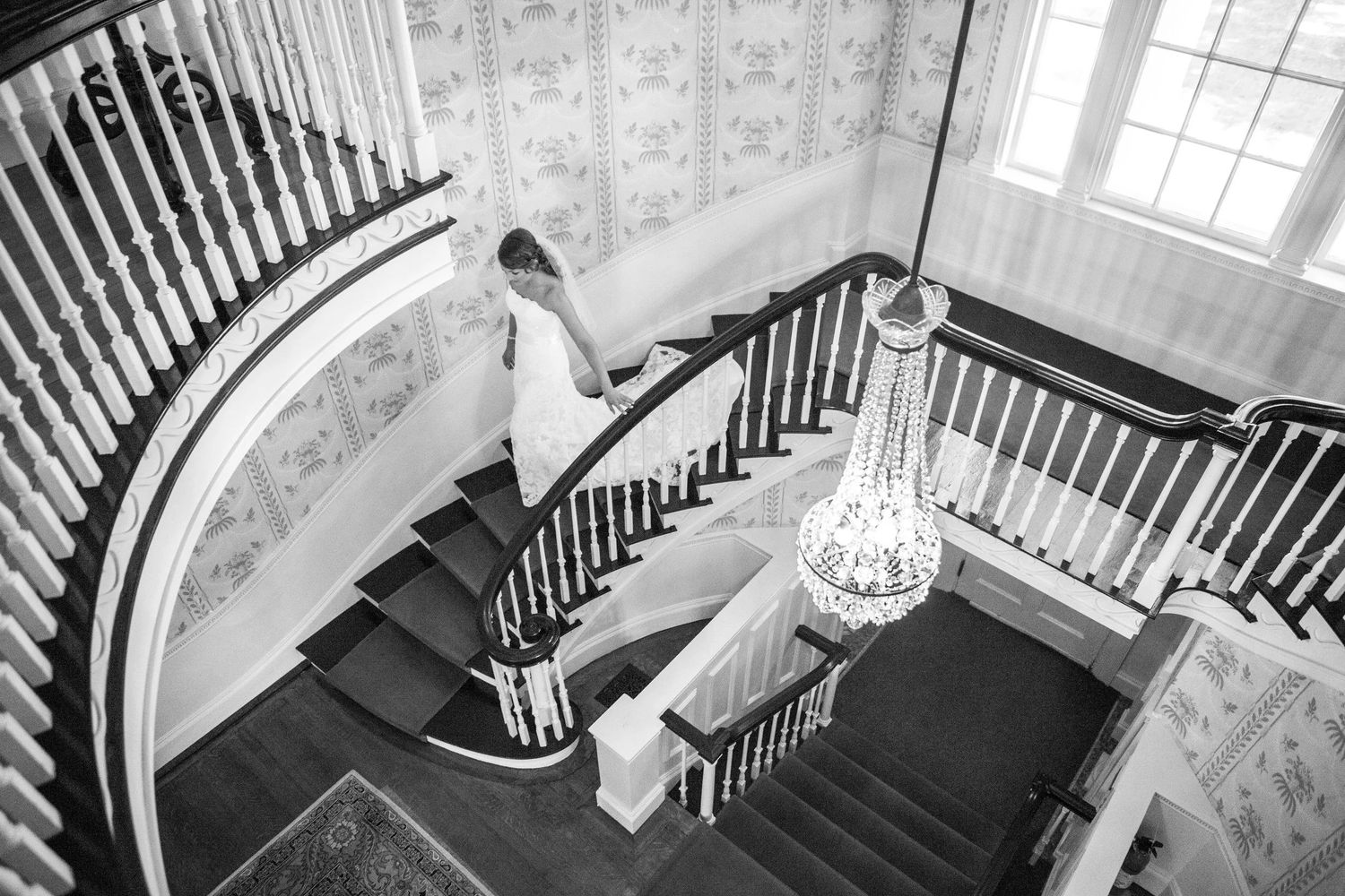 Black and white photo of a bride in a wedding gown descending an elegant curved staircase with a crystal chandelier.