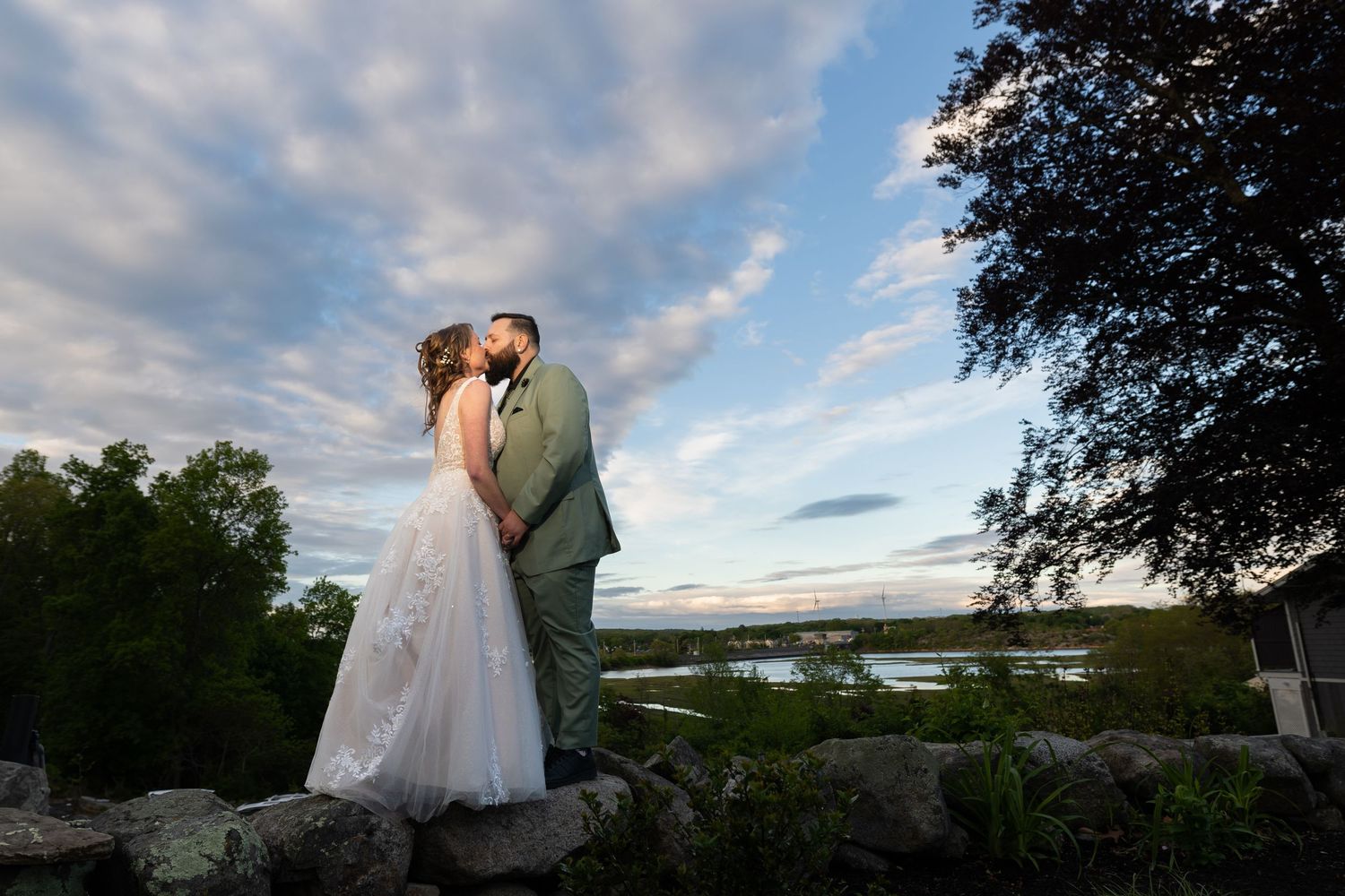 Bride and groom share a romantic kiss on stone wall at sunset with scenic water views and dramatic cloudy sky.