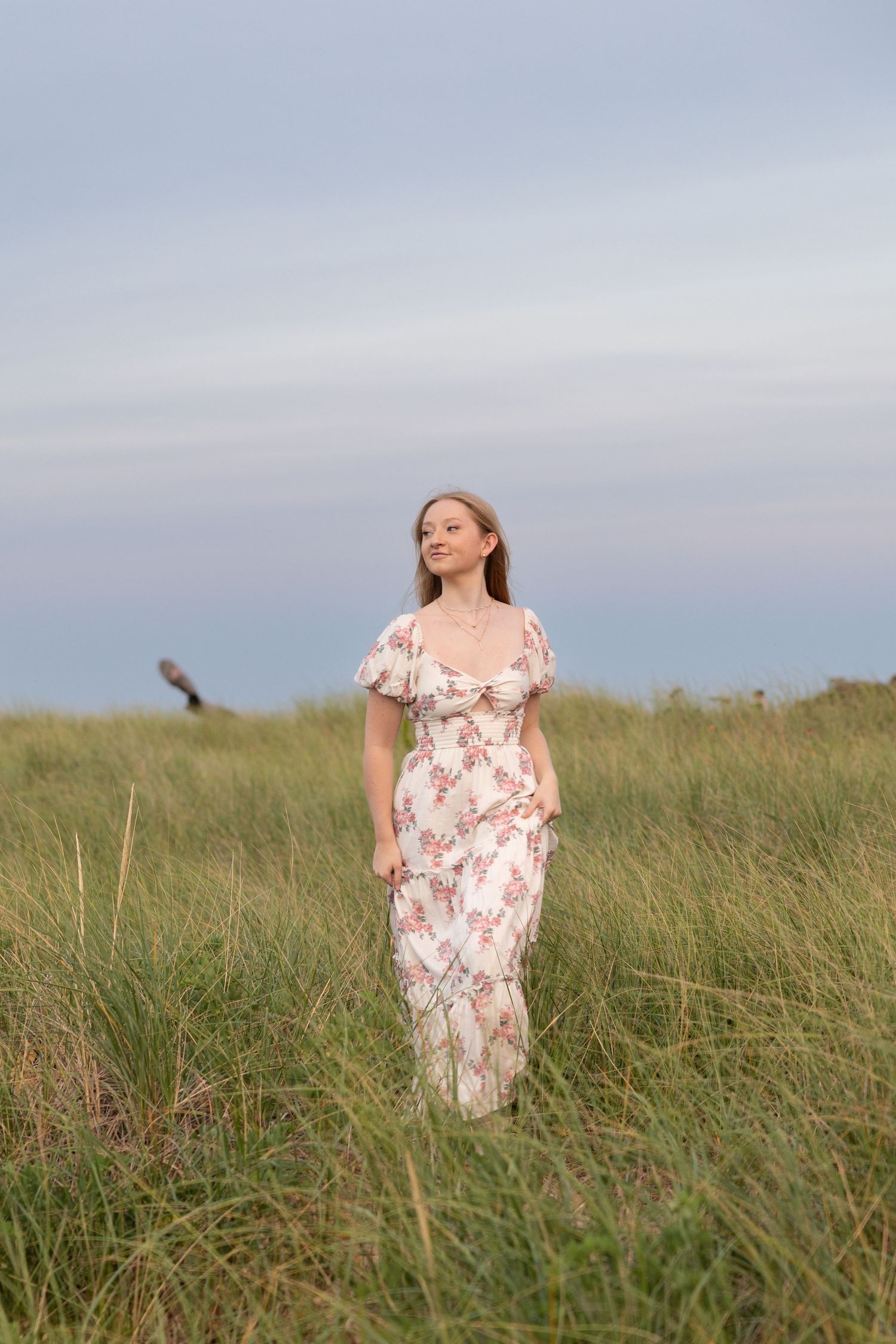 Young woman in floral maxi dress standing in tall green grass field under cloudy sky.