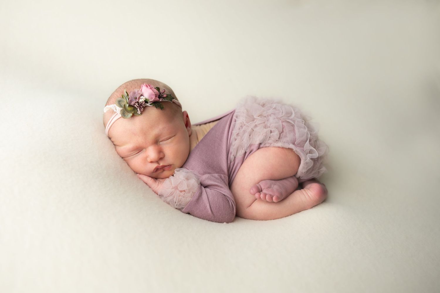 Sleeping newborn baby girl in lavender lace romper and floral headband posed on white backdrop.