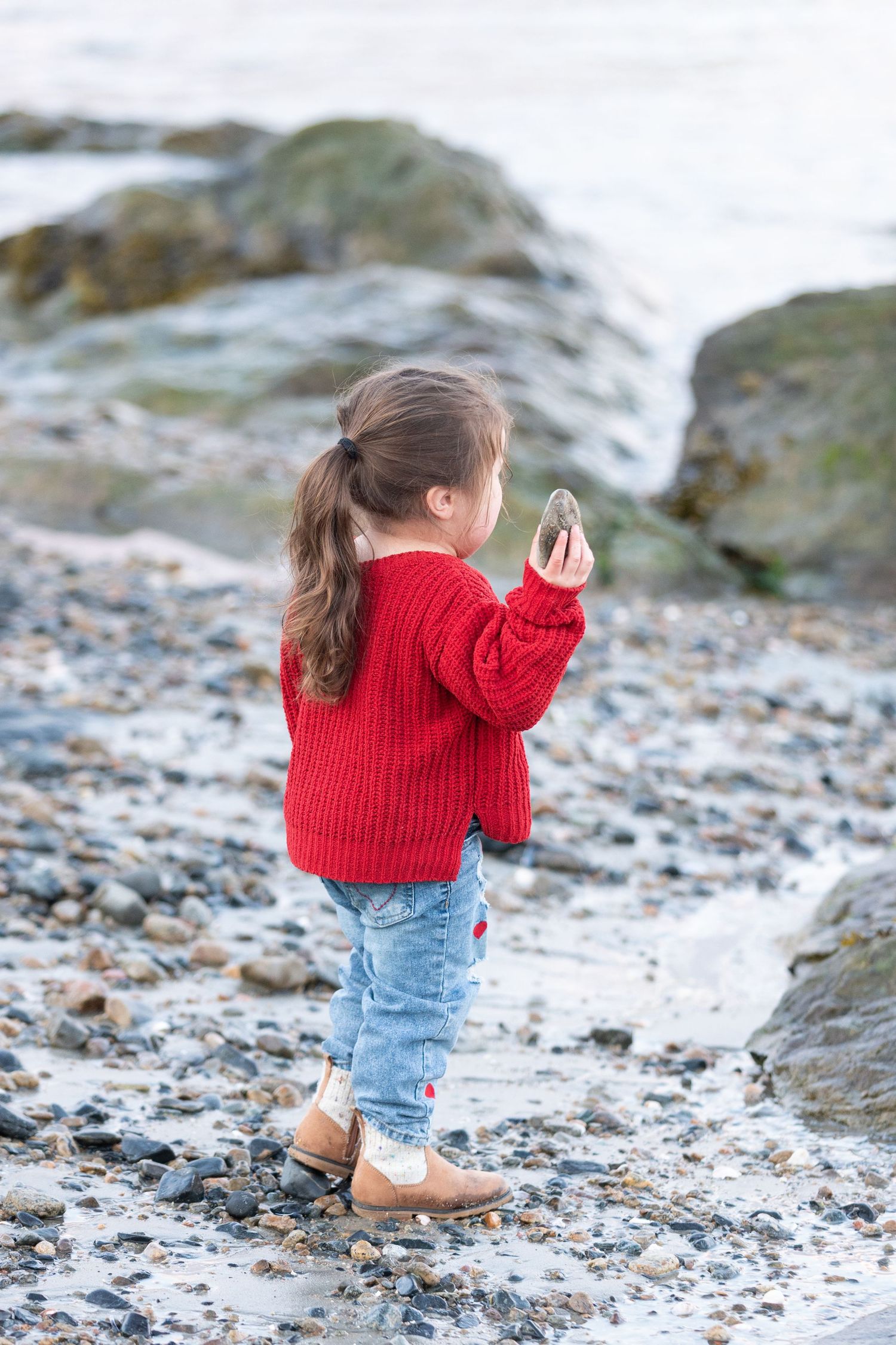 Young girl in red sweater and jeans holds a rock on a rocky beach shoreline.