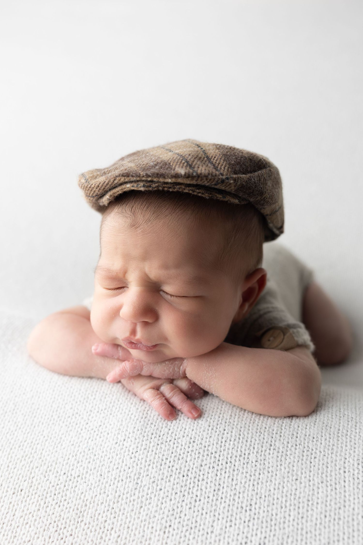 Newborn baby wearing a plaid flat cap rests chin on hands, sleeping peacefully on a white knit blanket.