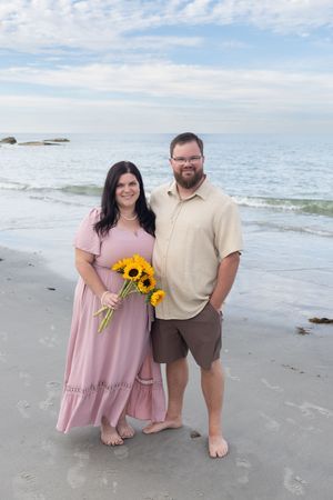 Couple posing on sandy beach, woman in pink maxi dress holding bright yellow sunflower bouquet.