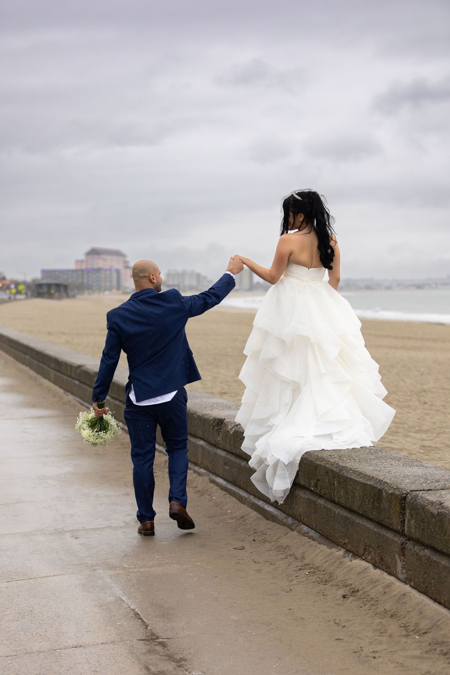 Bride in white ruffled gown balancing on seawall as groom in blue suit holds her hand on cloudy beach.