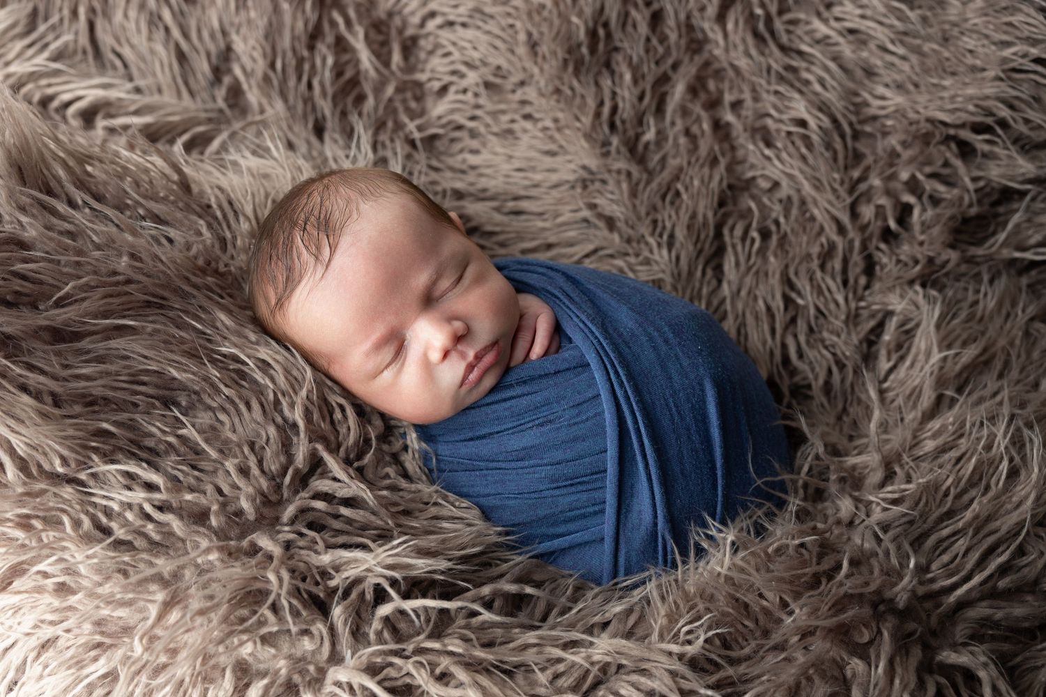 Peaceful newborn baby swaddled in blue wrap sleeping on fluffy gray fur blanket.