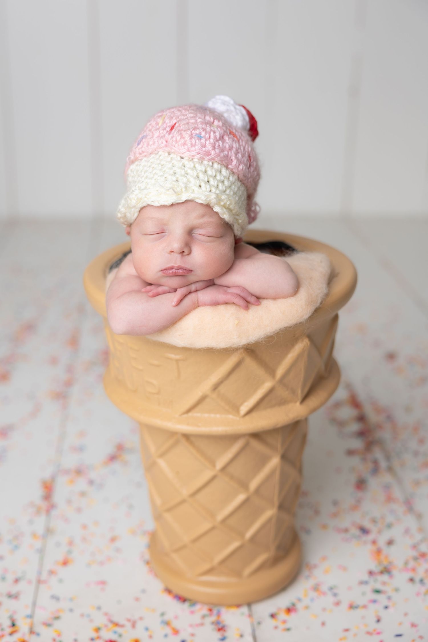 Sleeping newborn baby wearing a knit ice cream hat, nestled in a waffle cone prop with sprinkles on white floor.
