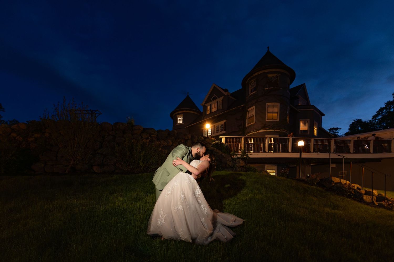 Bride and groom share romantic dip kiss at twilight in front of illuminated Victorian mansion wedding venue.