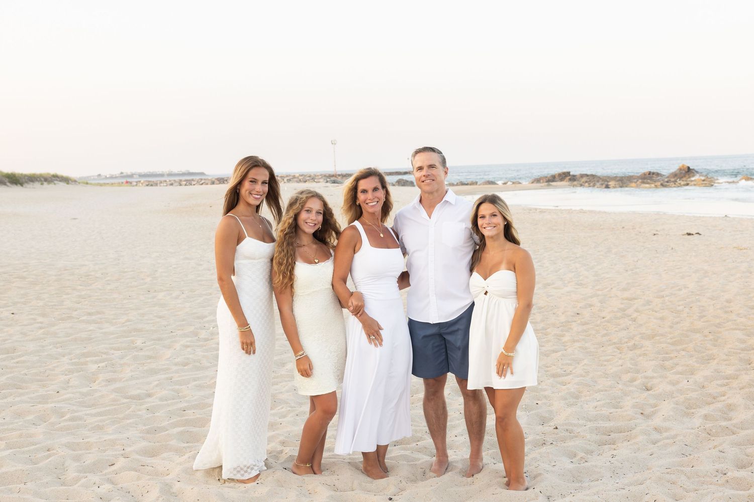 Family of five posing together on a sandy beach, all wearing white outfits, smiling during golden hour.
