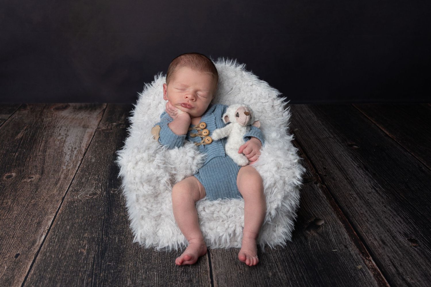 Newborn baby sleeping in a fluffy white bucket with a small stuffed animal, wearing a blue knit romper on rustic wood.