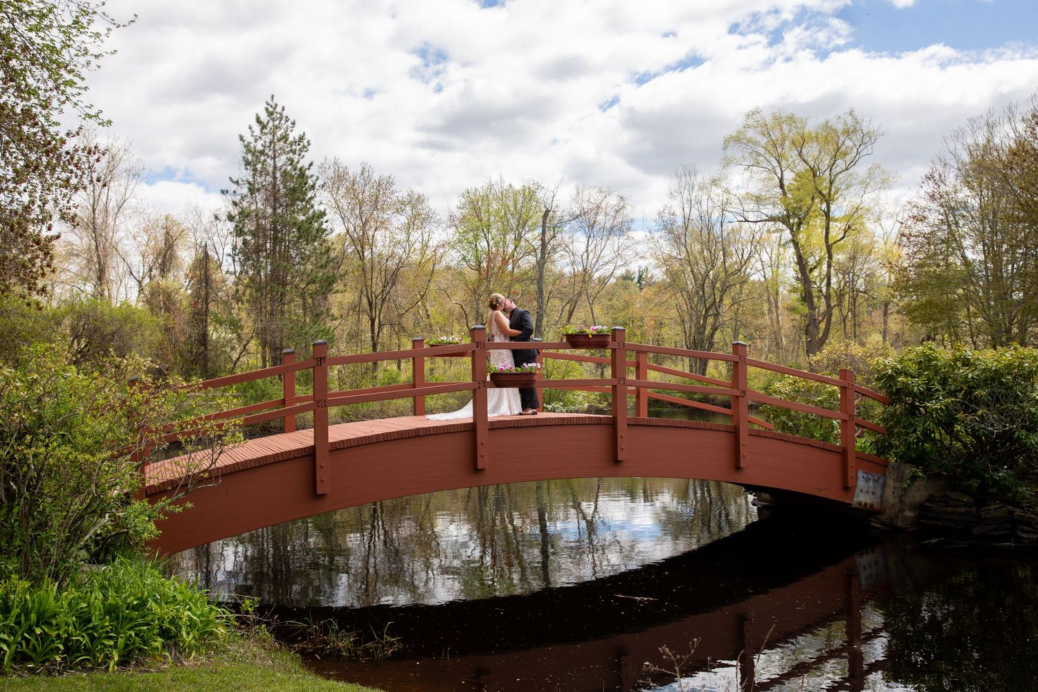 Couple embracing on a red arched bridge over a reflective pond surrounded by lush green trees on a cloudy day.