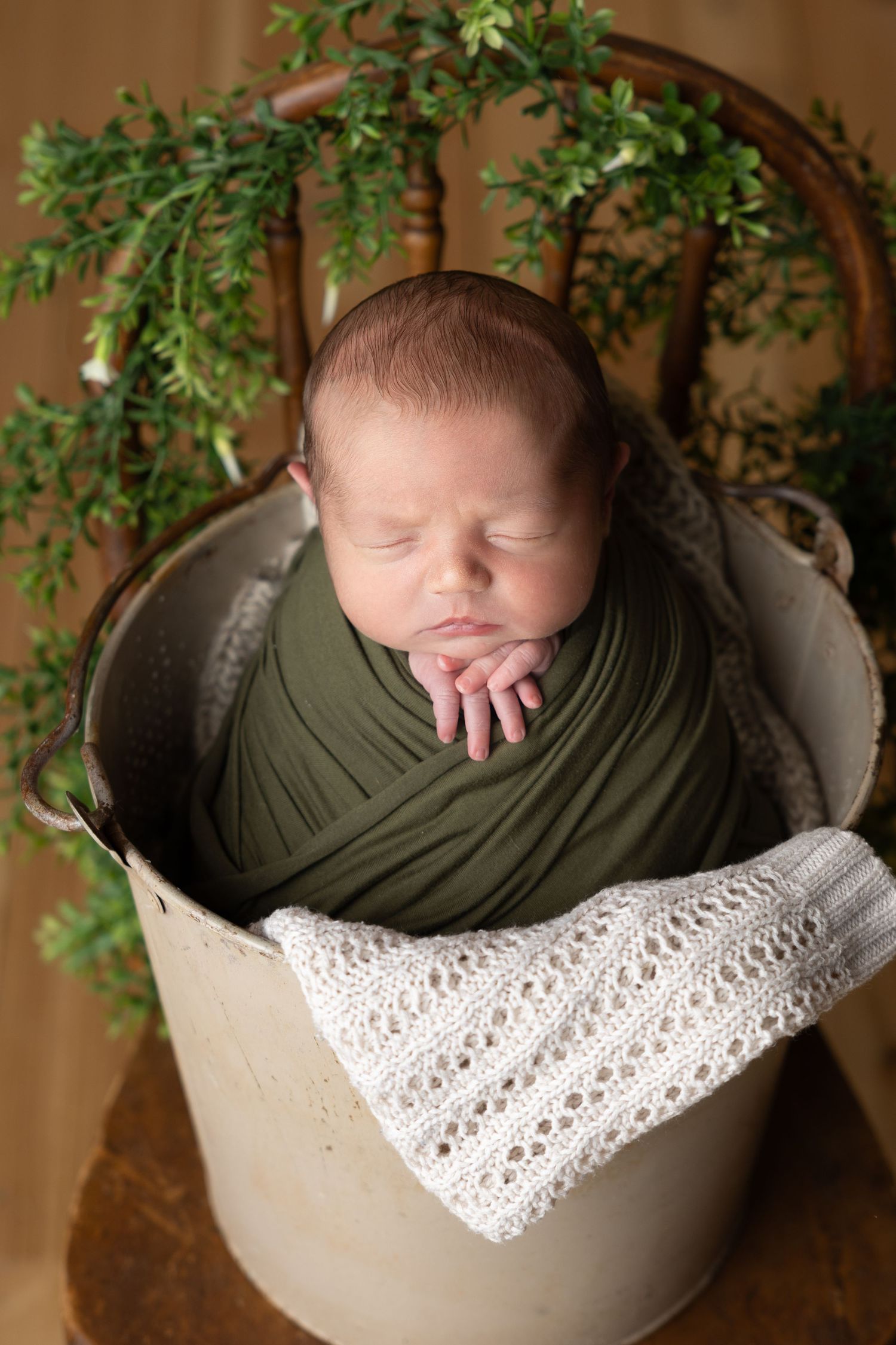 Newborn baby swaddled in green wrap sleeping peacefully inside a rustic metal bucket with lace blanket and greenery.