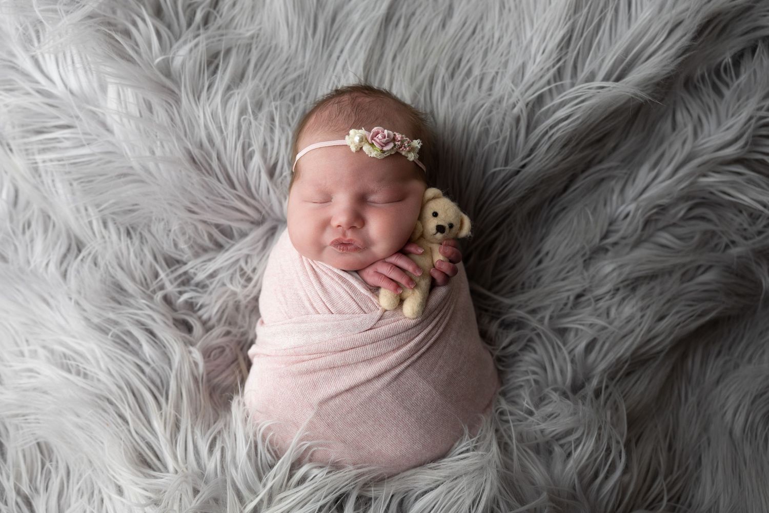 Newborn baby swaddled in pink wrap holding a tiny teddy bear, sleeping on a gray fluffy blanket with a floral headband.
