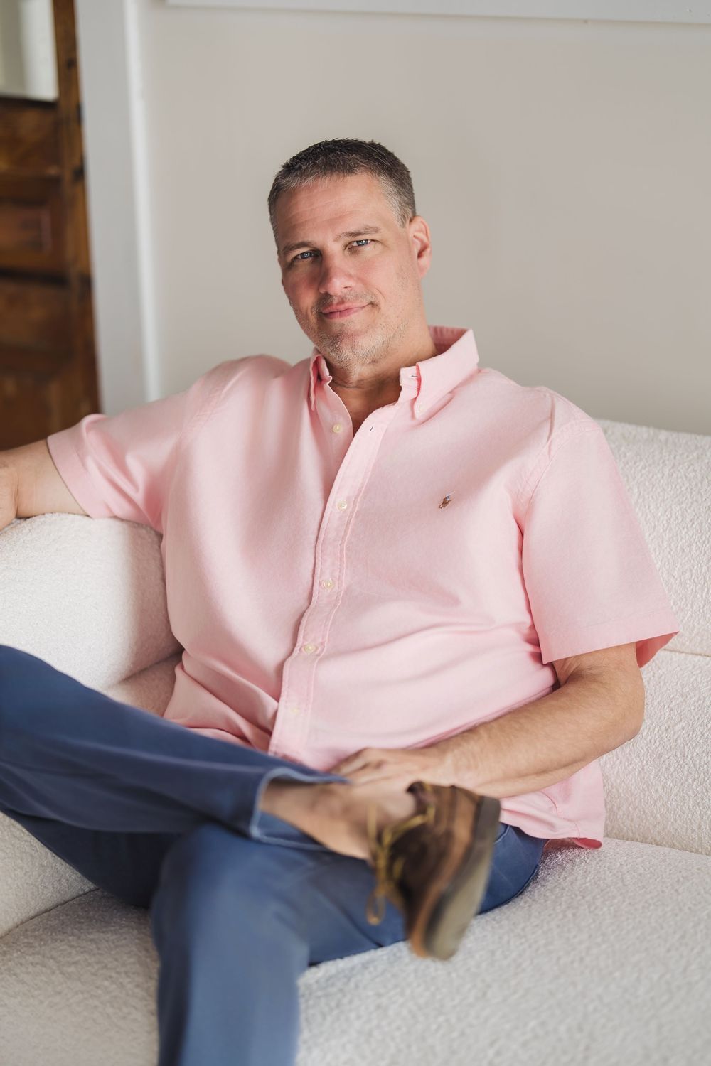 Relaxed middle-aged man in pink polo shirt sitting comfortably on white sofa at home.