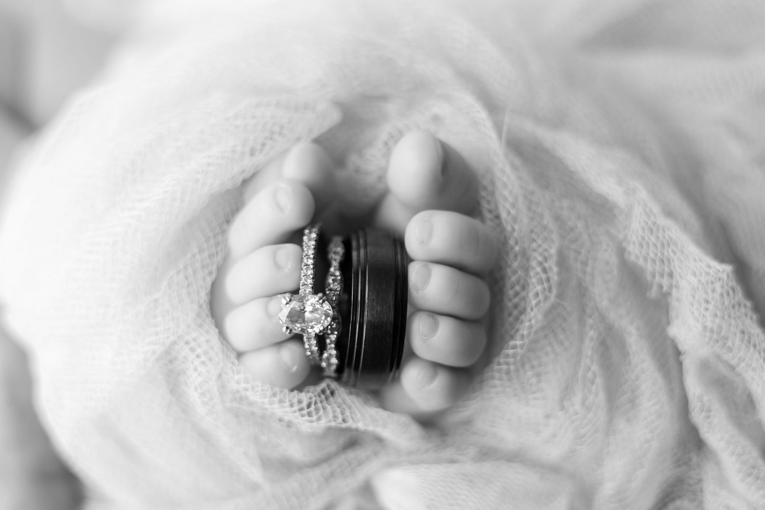 Black and white close-up of newborn baby fingers holding diamond engagement ring and wedding band.