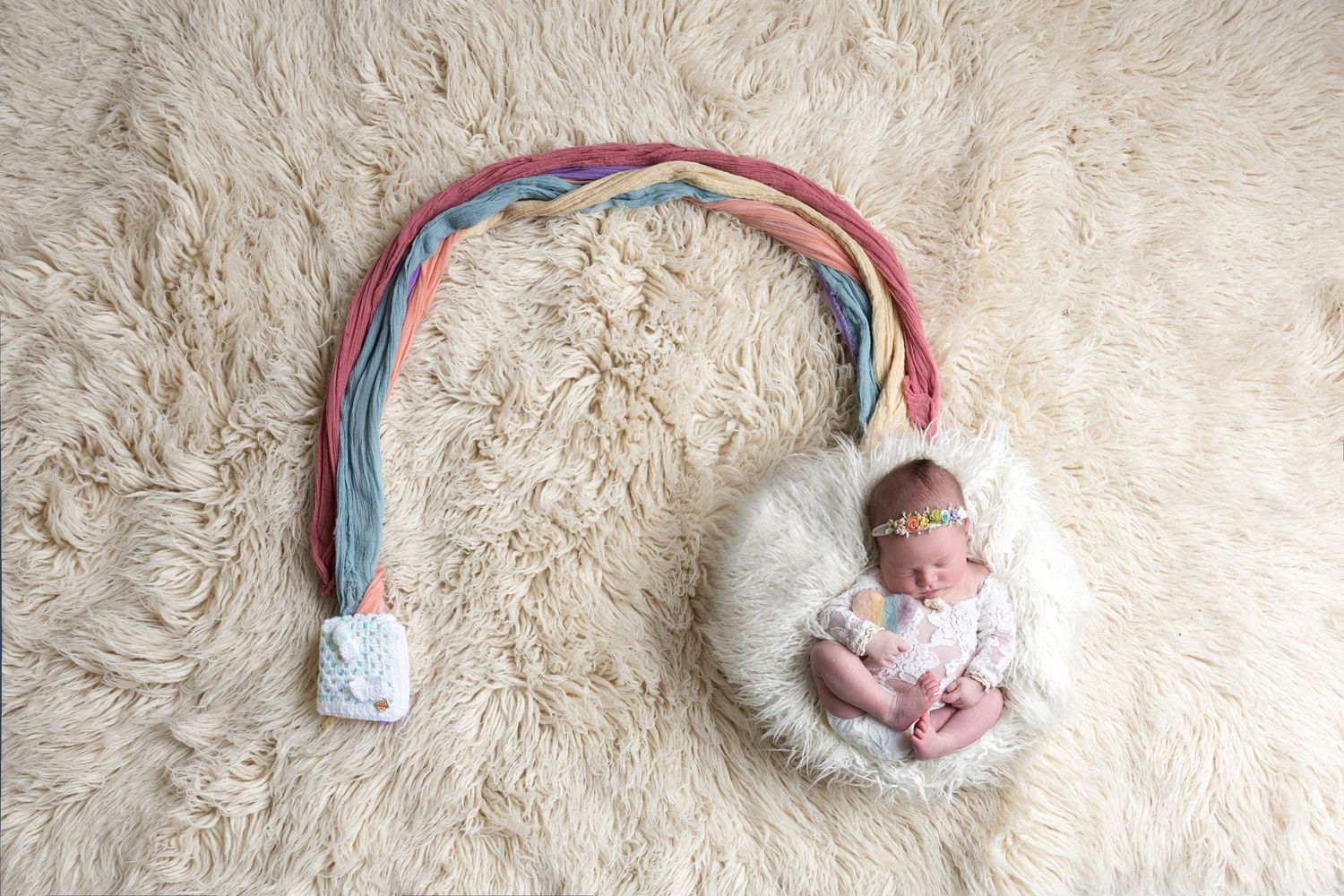 Newborn baby sleeping in a white bowl with a colorful rainbow prop on a fluffy cream rug.