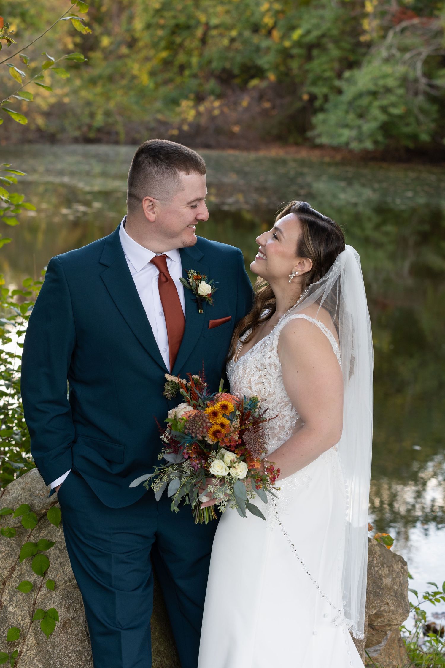 Bride and groom share a joyful moment by a pond, she holds autumn bouquet, he wears navy suit with red tie.