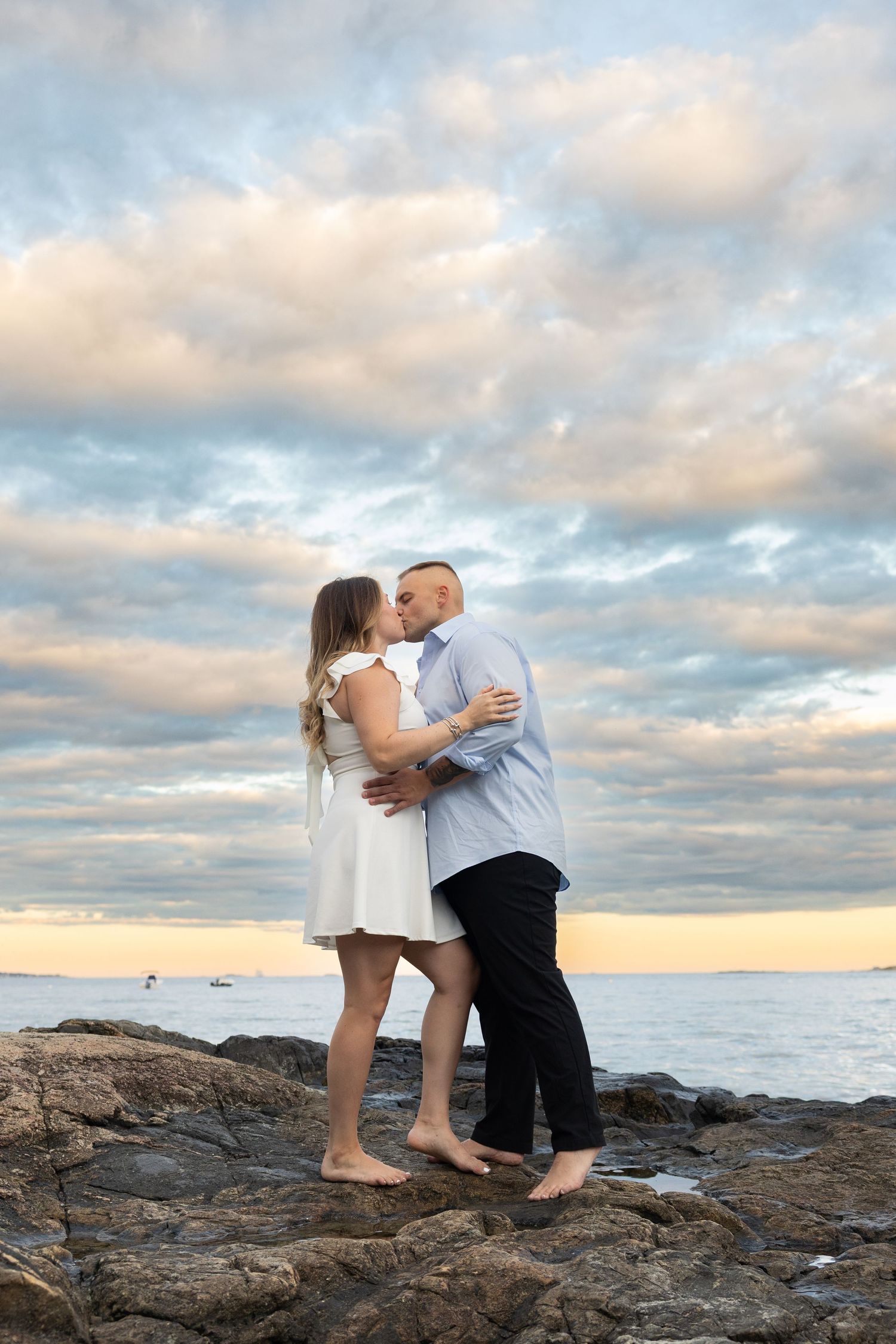 Couple kissing barefoot on rocky shore at sunset with dramatic cloudy sky and ocean in background.