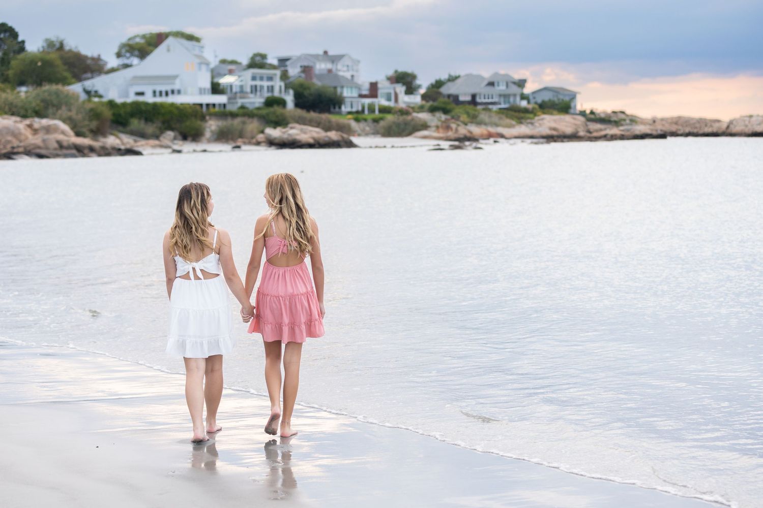 Two girls in dresses hold hands walking along a sandy beach with coastal homes in the background.
