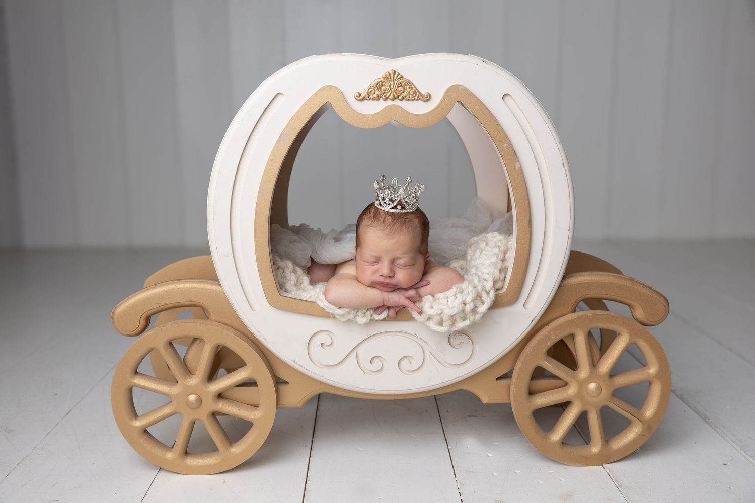 Newborn baby sleeping in a white and gold Cinderella carriage prop wearing a tiny princess crown.