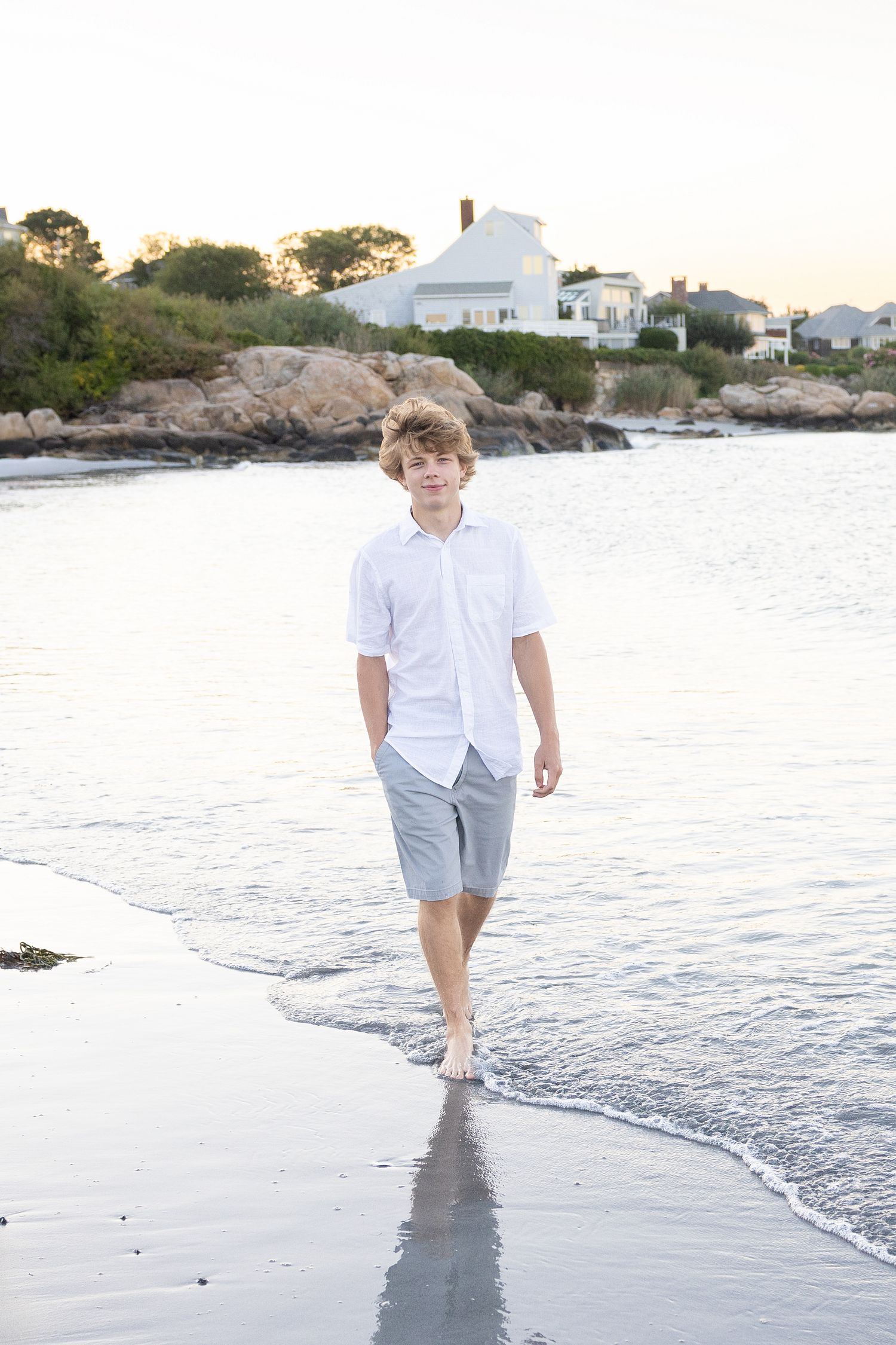 A teenage boy in a white shirt and gray shorts walks barefoot along a sandy beach at sunset.