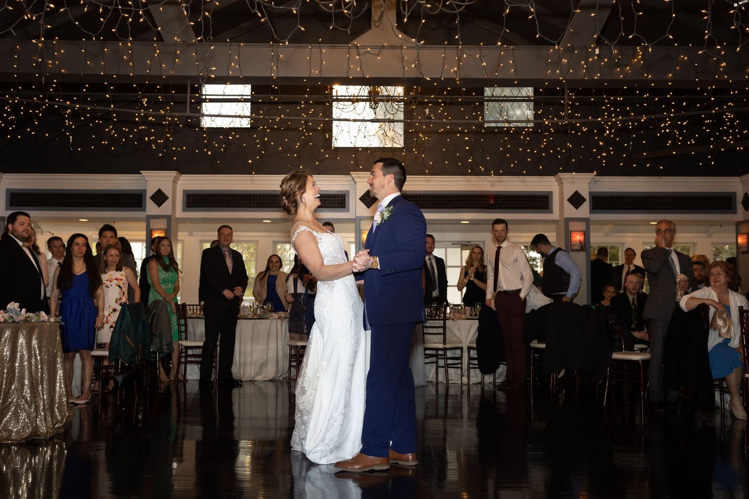 Bride and groom share their first dance at elegant wedding reception with twinkling lights and guests watching.