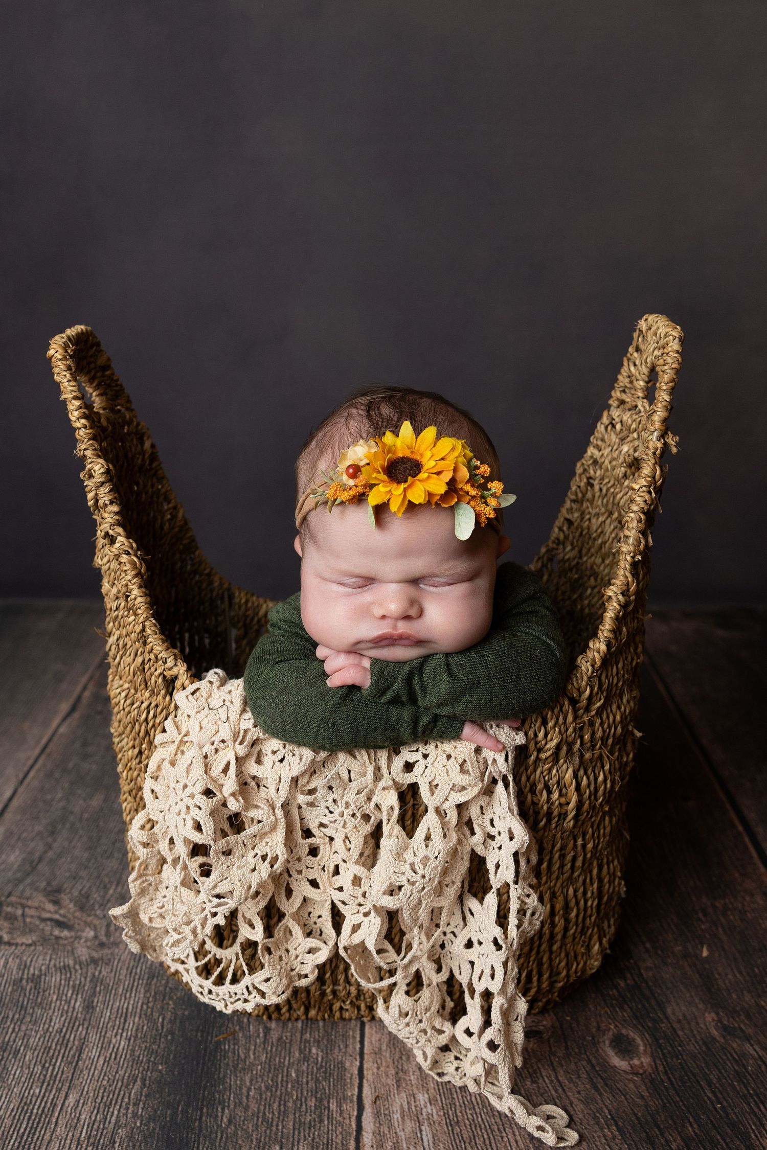 Sleeping newborn baby girl in a woven basket wearing a sunflower headband, nestled in green and cream lace blankets.