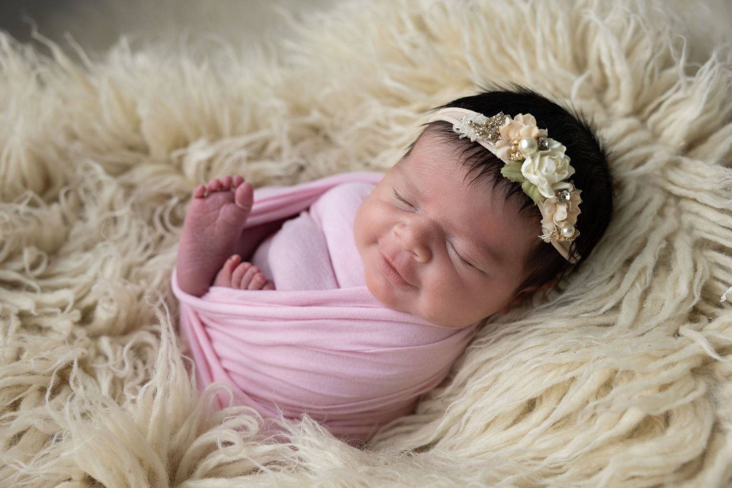 Sleeping newborn baby girl wrapped in pink swaddle with floral headband on fluffy cream blanket.