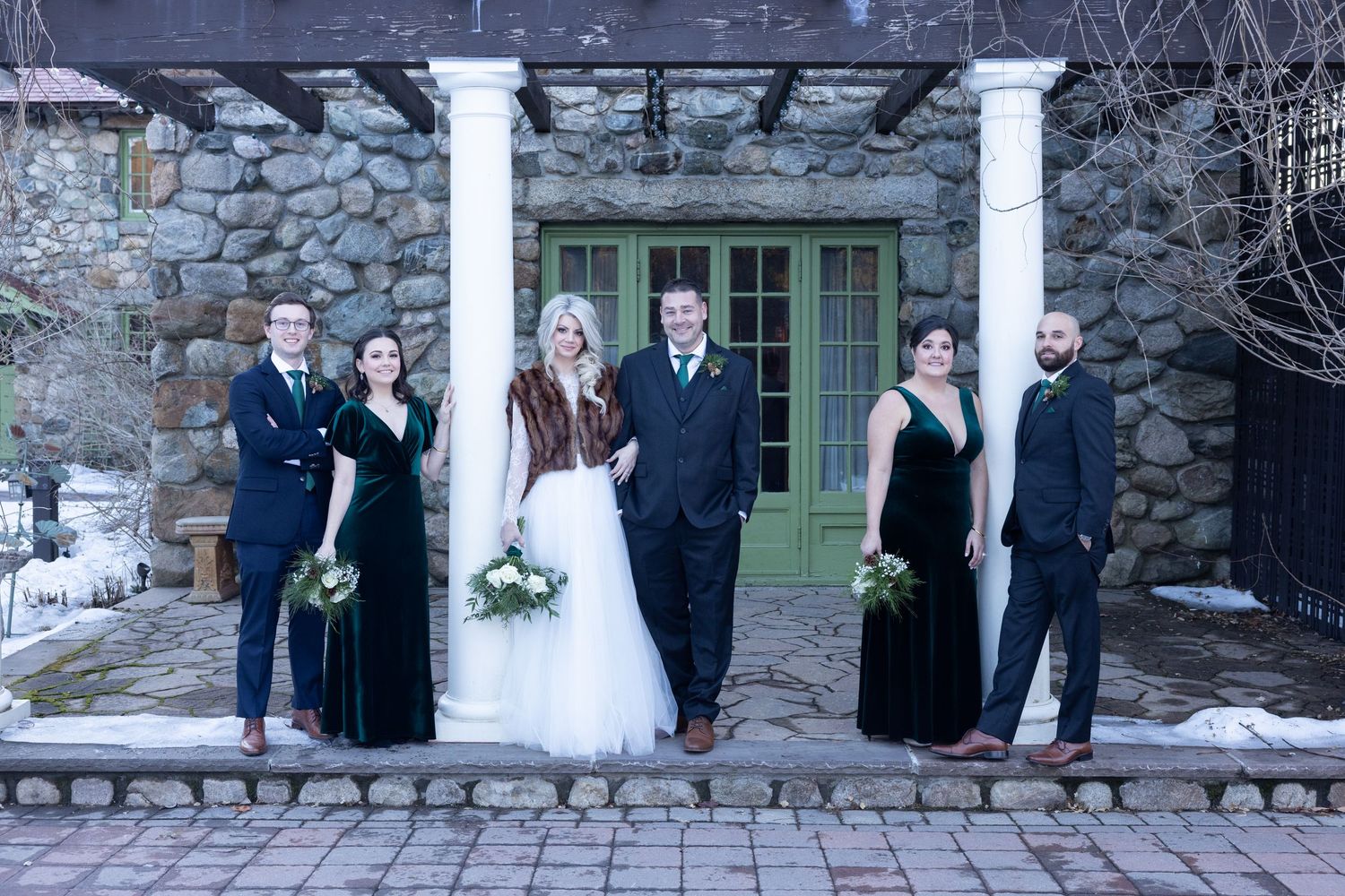 Wedding party in winter, bride and groom with bridesmaids in velvet gowns and groomsmen before stone building.
