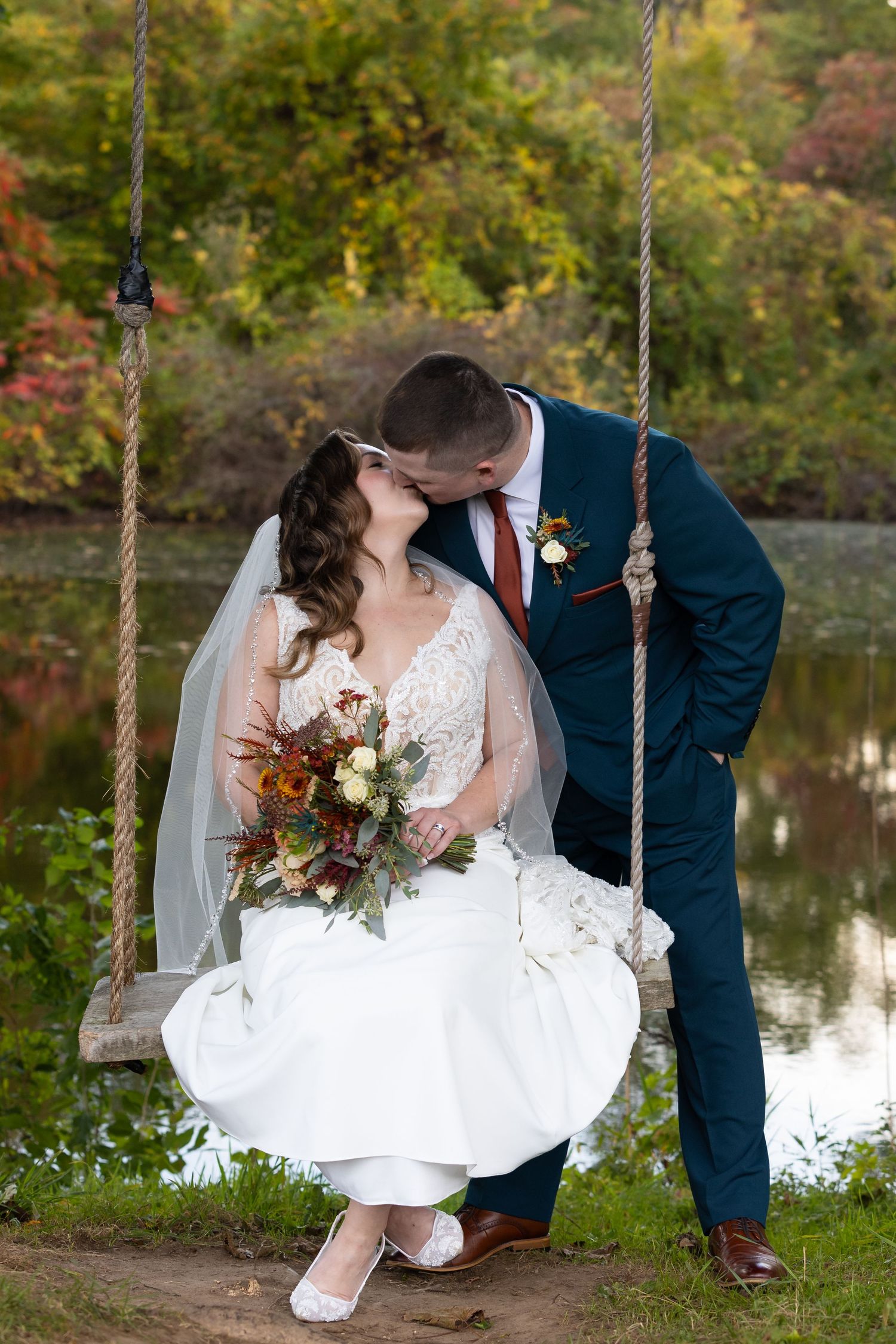 Bride and groom share a kiss on a rope swing near a pond surrounded by colorful autumn foliage.