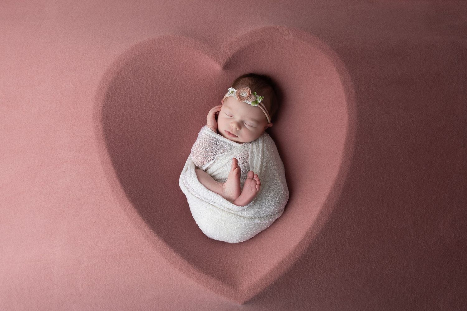 Newborn baby swaddled in white wrap sleeping peacefully inside a heart-shaped cutout on a pink background.