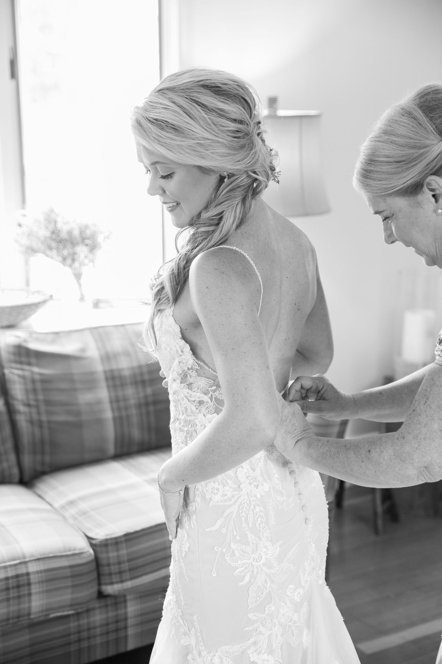 Bride in lace wedding gown being buttoned up by bridesmaid in elegant black and white photo.