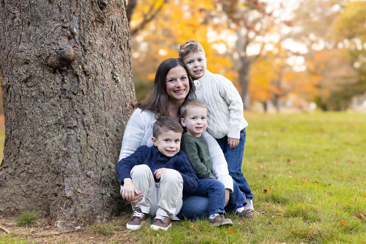A mother poses with her three young sons beside a large tree in a park during autumn.