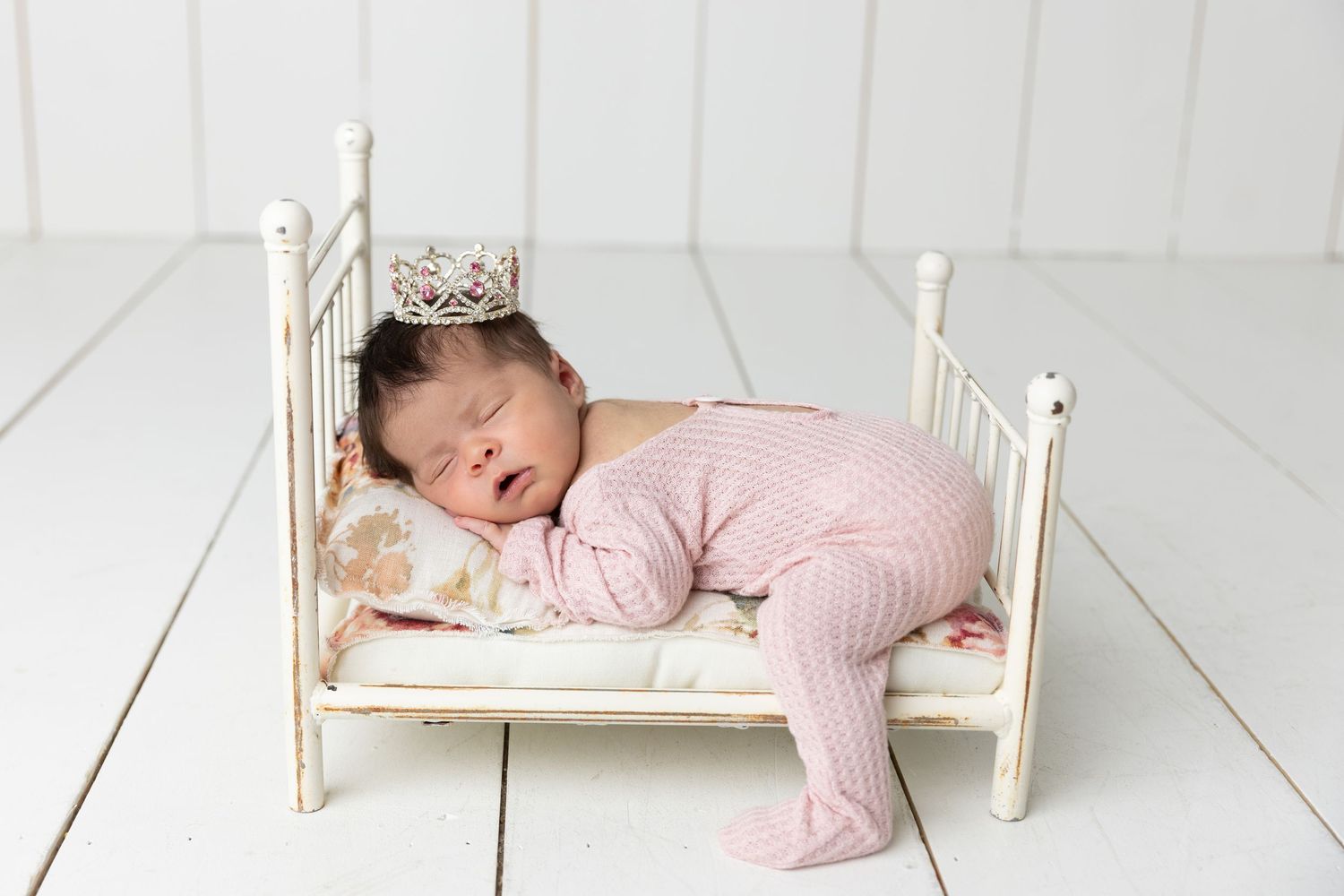 Newborn baby in pink onesie and tiny crown sleeping peacefully on a miniature white vintage bed.