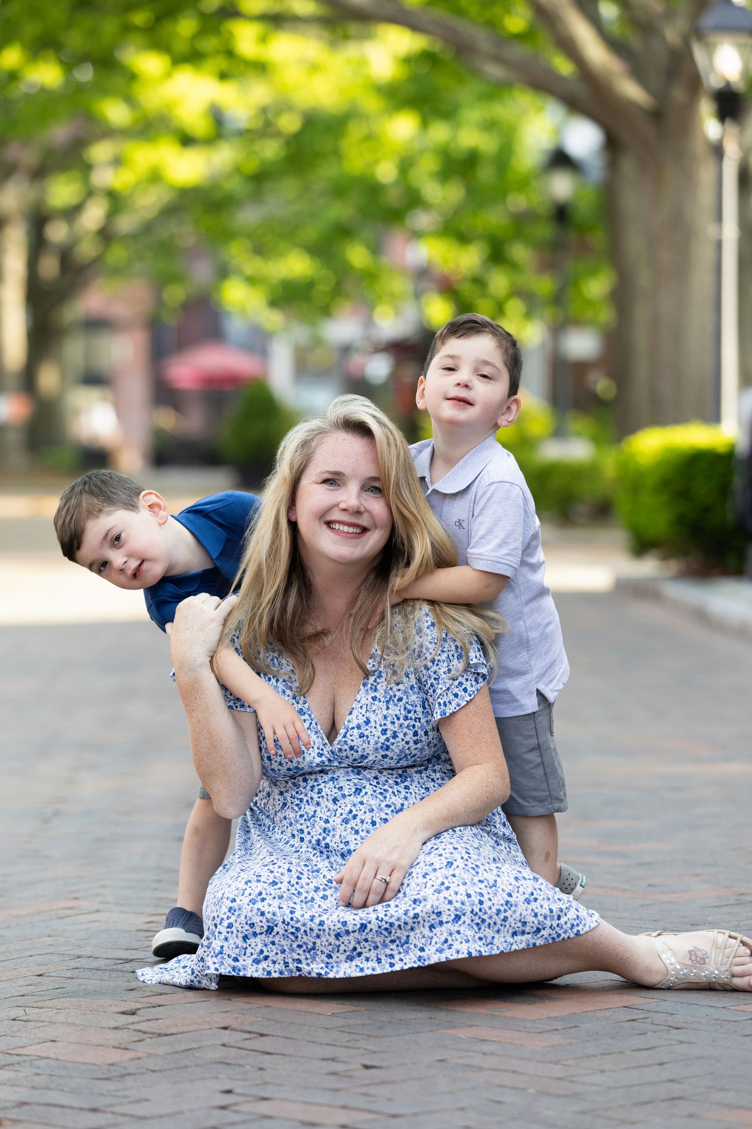 A smiling blonde woman in a floral dress sits on a brick path with two young boys hugging her outdoors.
