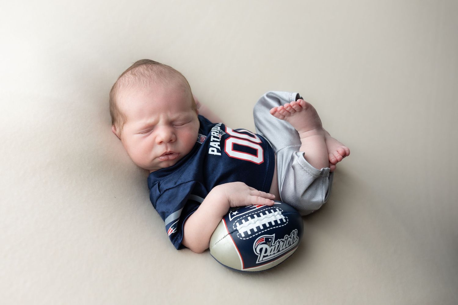 Newborn baby sleeping peacefully in a Patriots football jersey, cradling a miniature Patriots football.