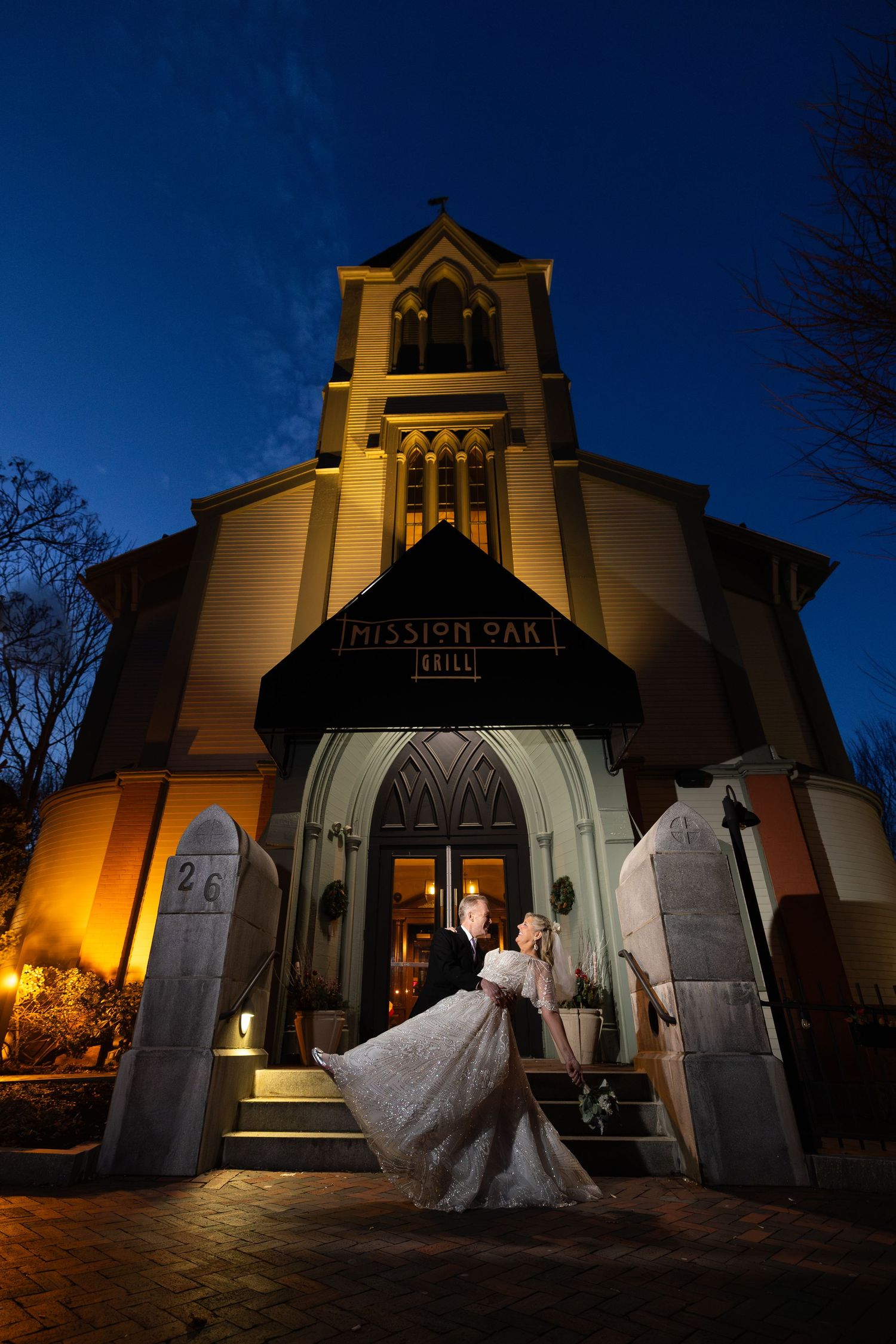 Couple dancing in wedding attire outside the illuminated Mission Oak Grill at twilight with a deep blue night sky.