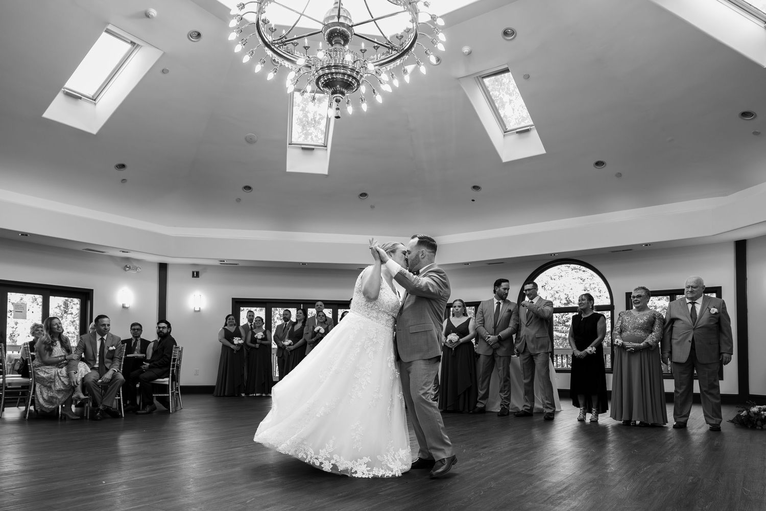 Bride and groom share their first dance in elegant ballroom while wedding guests watch, captured in black and white.