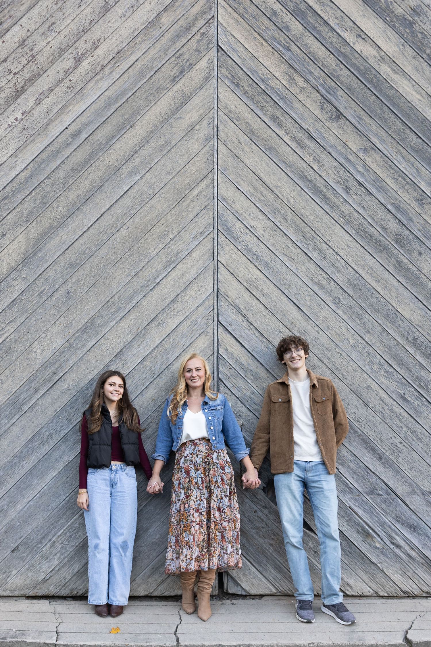 Three smiling people holding hands in front of a rustic wooden chevron wall, wearing casual fall outfits.