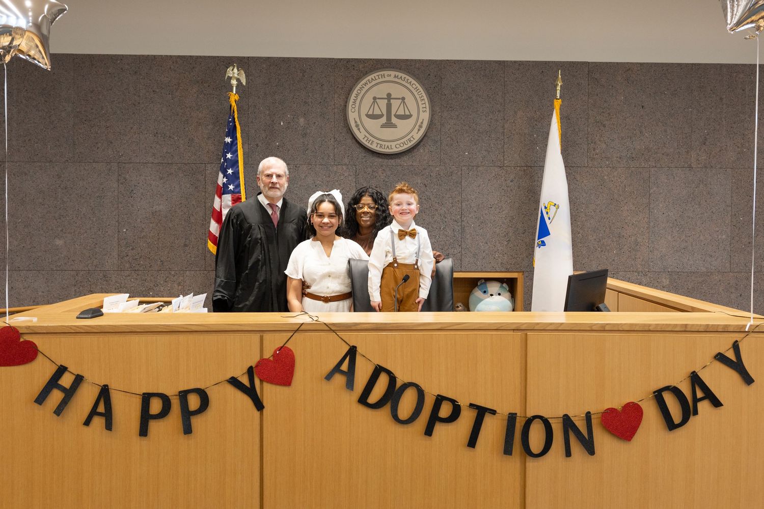 Family celebrates Happy Adoption Day in courtroom with judge, banner, and American flag backdrop.