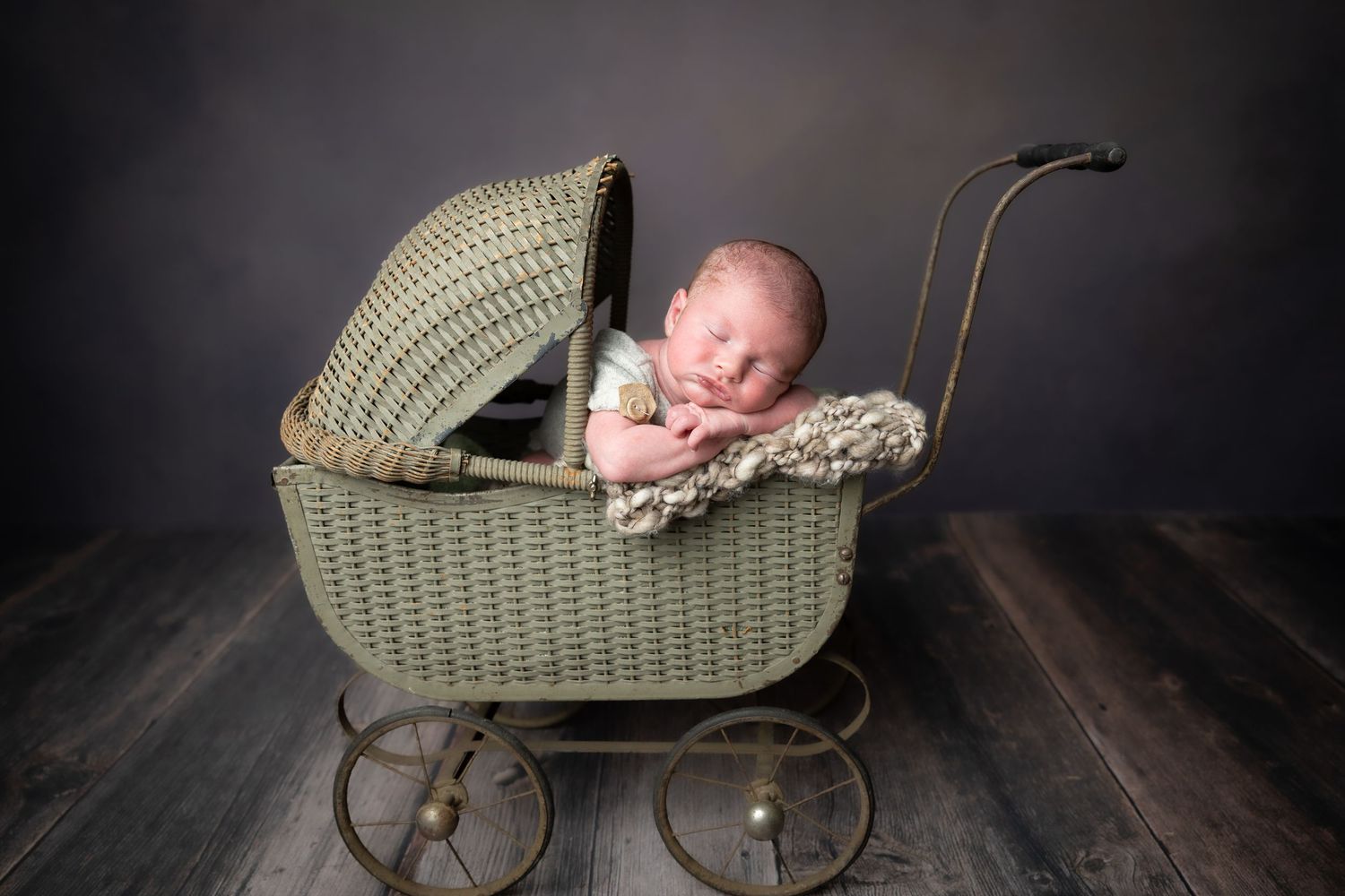 Newborn baby sleeping peacefully in a vintage green wicker pram on a rustic wood floor.