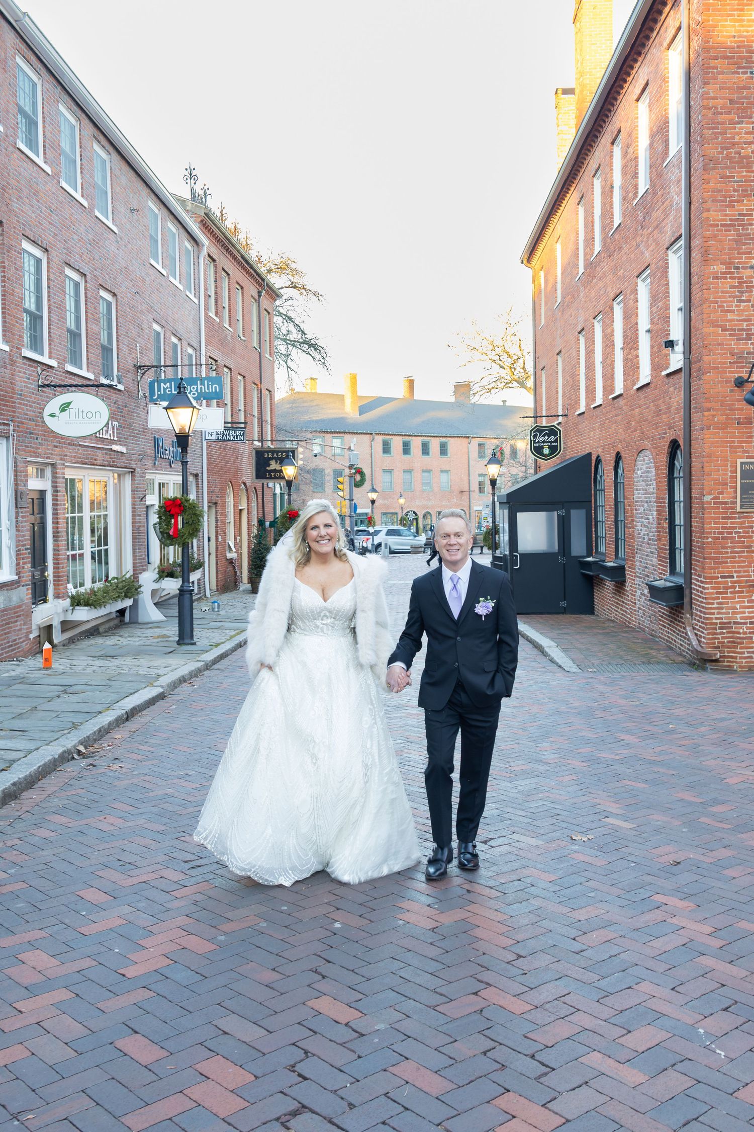 Bride and groom walk hand-in-hand on a historic brick street lined with red buildings during their winter wedding.