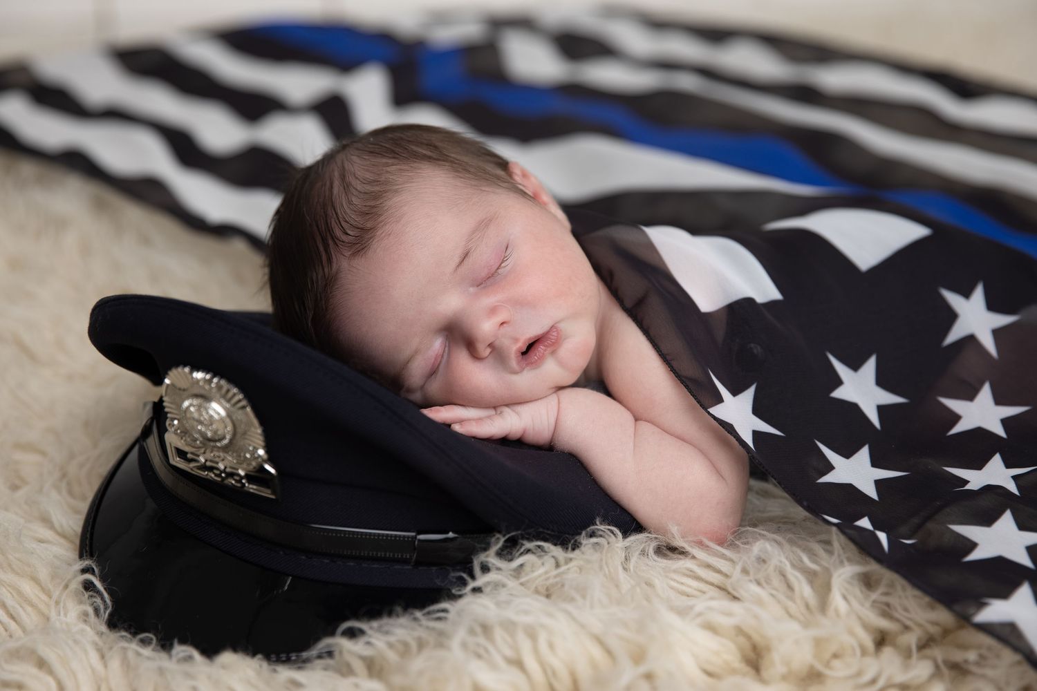 Newborn baby sleeping peacefully on a police officer hat, wrapped in a thin blue line American flag blanket.